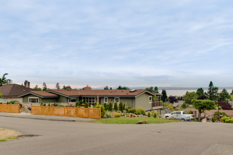 Single family rancher-style home in the Broadview/Bitter Lake neighborhood of Seattle, Washington. House has mature landscape on sloping yard and view of water.