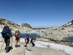 Team Diva Real Estate - Seattle - National Hiking Day - photo of group of people with backpacks hiking down mountain towards glacier lake