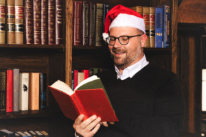 Roy Powell reading a book in front of a bookshelf while wearing a santa hat